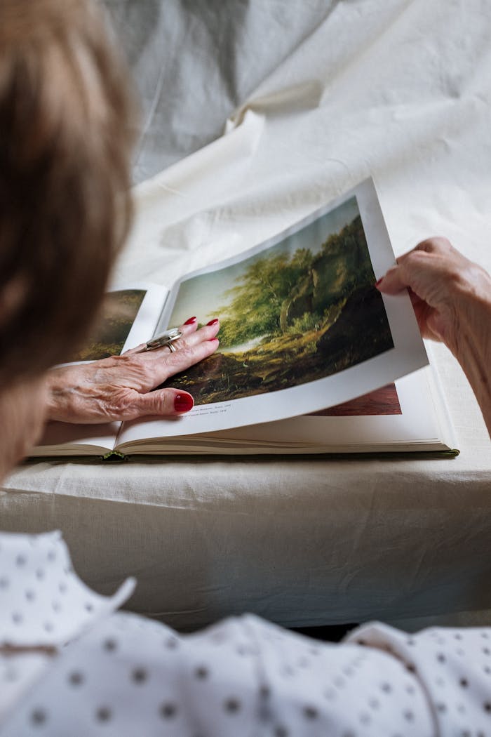 embark An elderly woman turns pages of a landscape art book at home, exploring detailed illustrations.