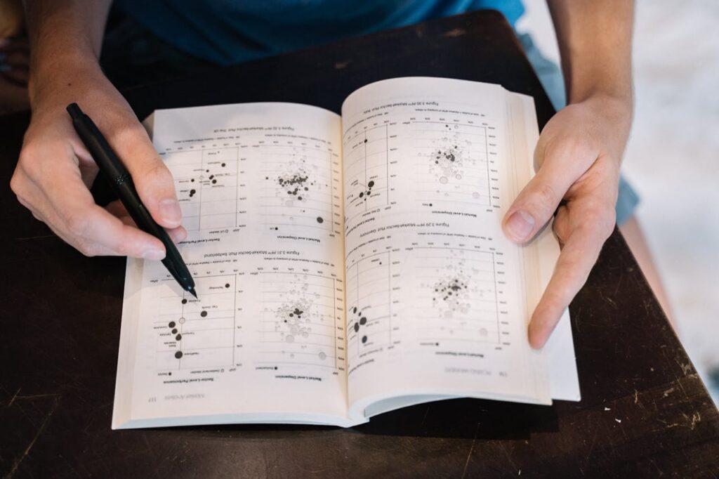 pexels photo 5527519 Person analyzing data charts in a book using a pen, close-up perspective.